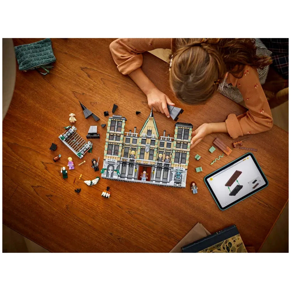 A child builds the LEGO Harry Potter Malfoy Manor set on a wooden table, surrounded by scattered pieces. An instruction tablet and a plush toy are visible.