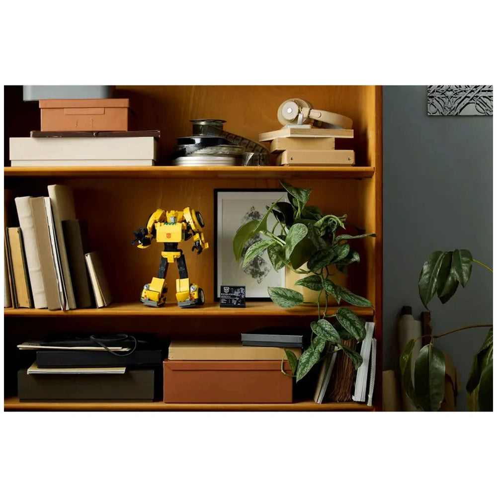 LEGO Icons Bumblebee construction set displayed on a wooden shelf surrounded by books, boxes, and a green plant.