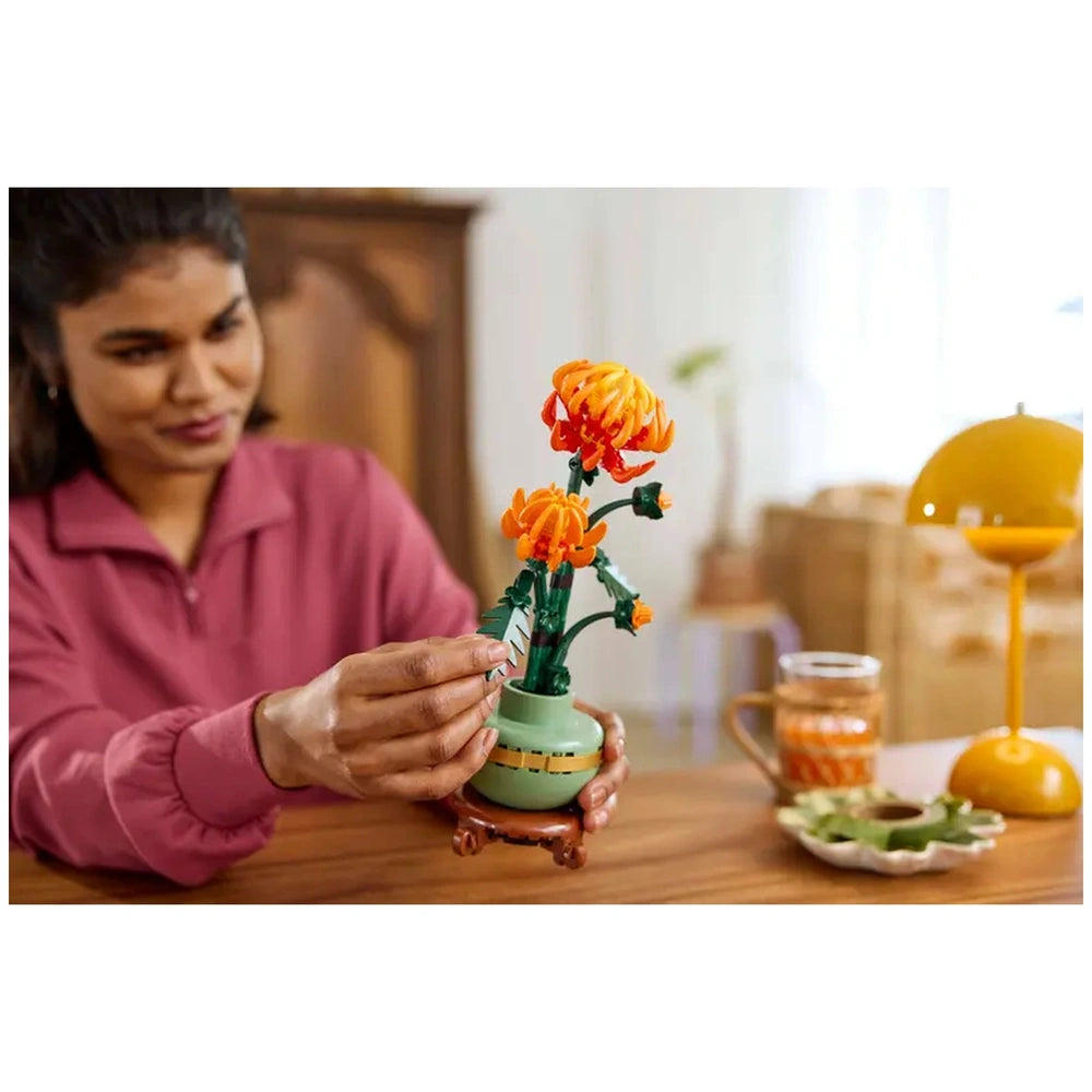 A person dressed in a pink sweater carefully adjusts an orange LEGO® chrysanthemum flower arrangement in a pastel green pot. The vibrant display features multiple blooms and lush green leaves, set on a wooden table with a vintage lamp and a glass mug nearby.