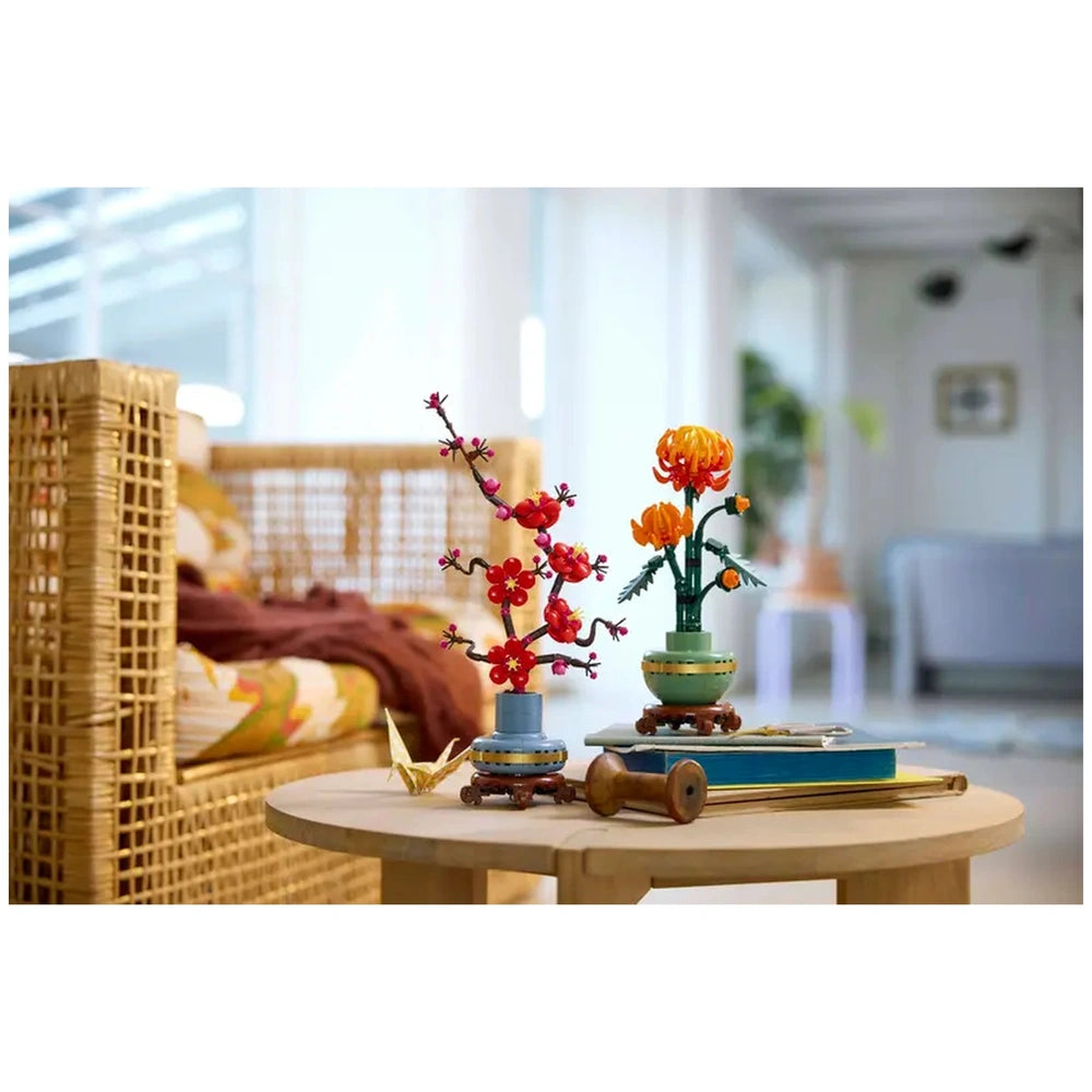 Vibrant LEGO® Icons Chrysanthemum set showcasing a bright orange flower, a blossoming chrysanthemum, and a branch of red flowers, elegantly arranged in decorative pots atop a wooden table, with a cozy living room background.