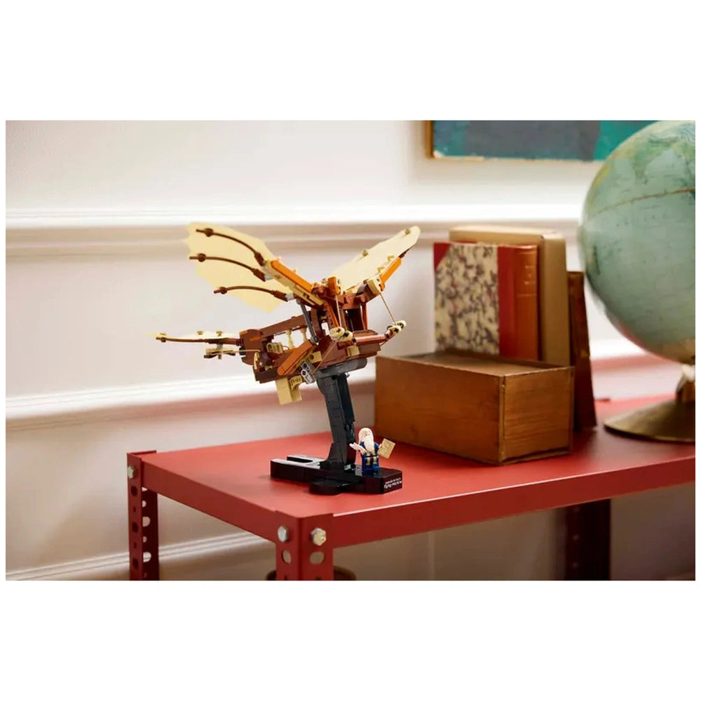 LEGO® Icons Leonardo da Vinci's Flying Machine model with flappable wings, displayed on a red table alongside books and a globe.
