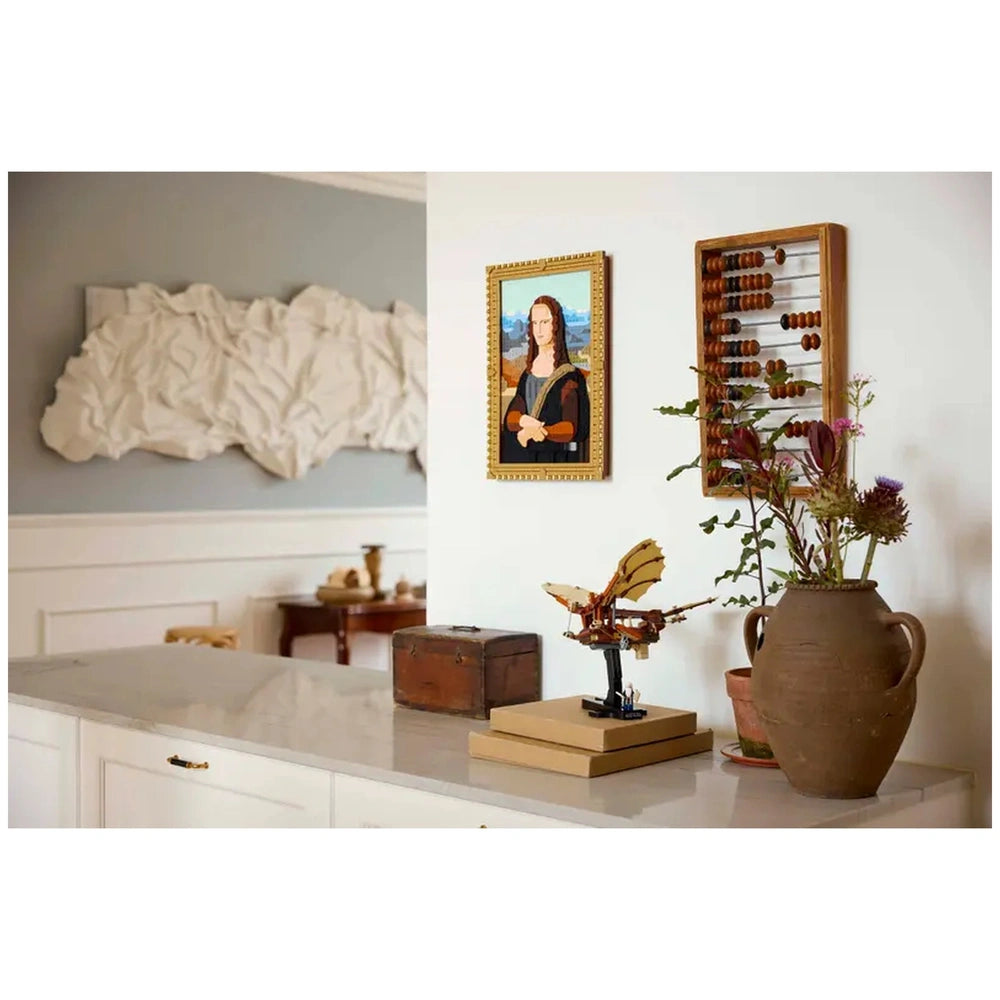 LEGO® Icons Leonardo da Vinci's Flying Machine displayed on a marble surface beside a vintage wooden box and potted flowers. A framed portrait, inspired by the Mona Lisa, hangs on the wall, next to an abacus.