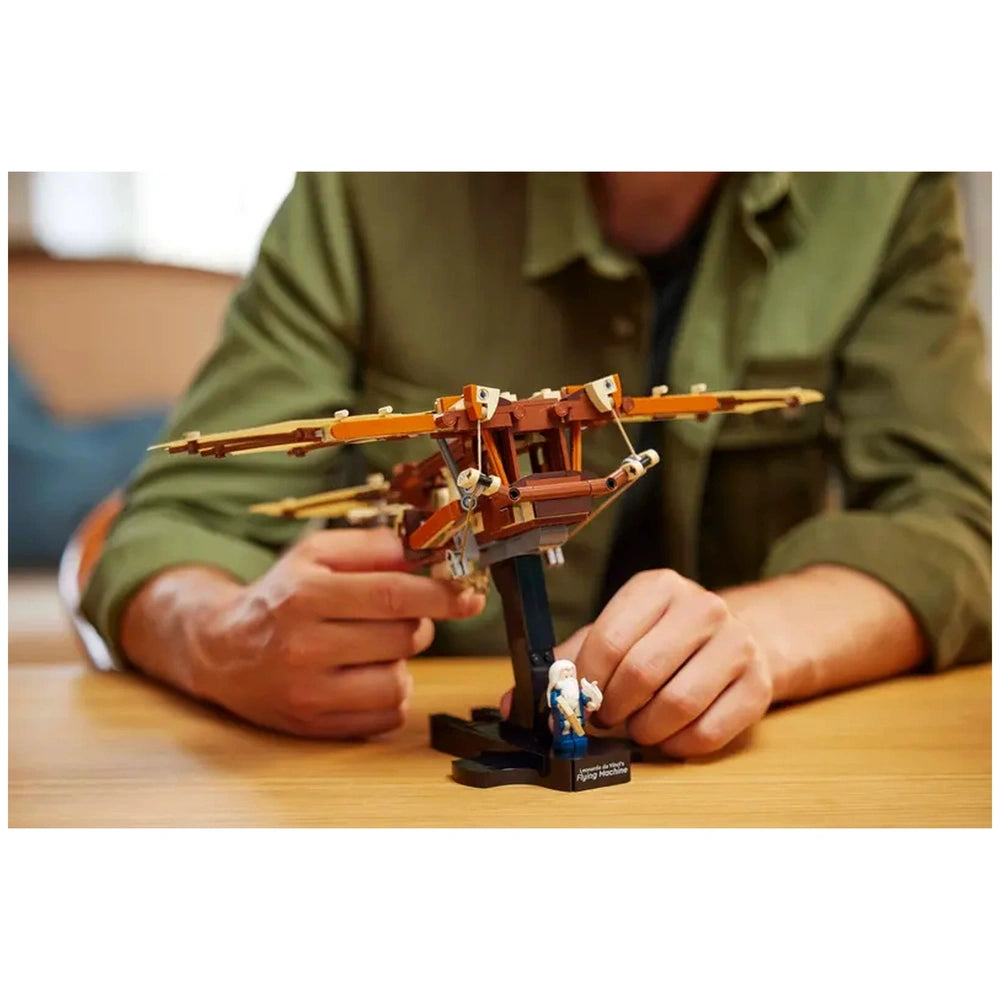LEGO® Icons Leonardo da Vinci's Flying Machine construction set, featuring a detailed model with flappable wings, displayed on a table. A person in a green shirt adjusts the model, with a miniature figure of Leonardo da Vinci nearby.