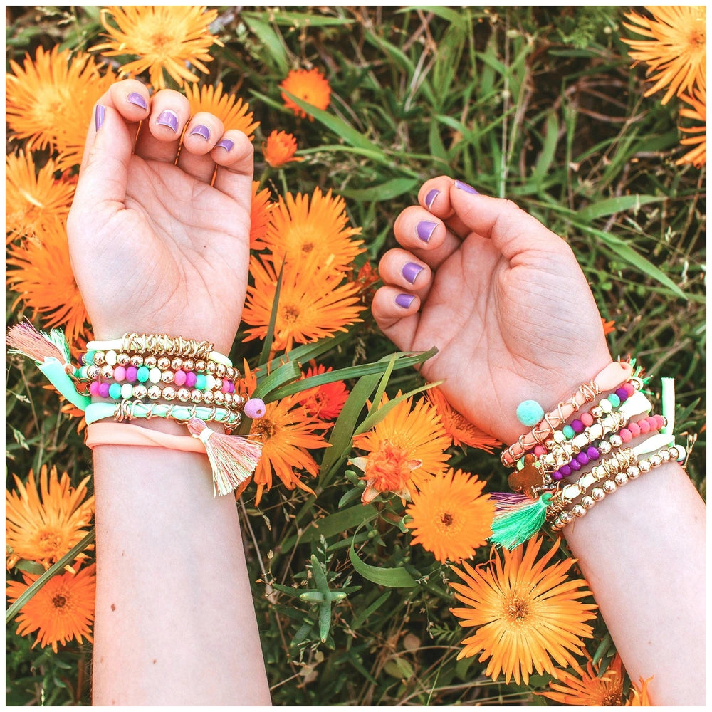 Neo-Brite chains and charms adorn two hands amidst vibrant orange and yellow flowers in a close-up lifestyle scene.