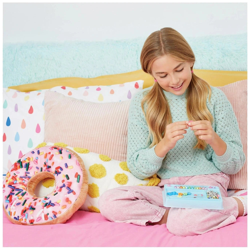 Young girl in a light blue sweater holds a blue container on a pink couch, with a colorful polka dot pillow and a pink donut 
