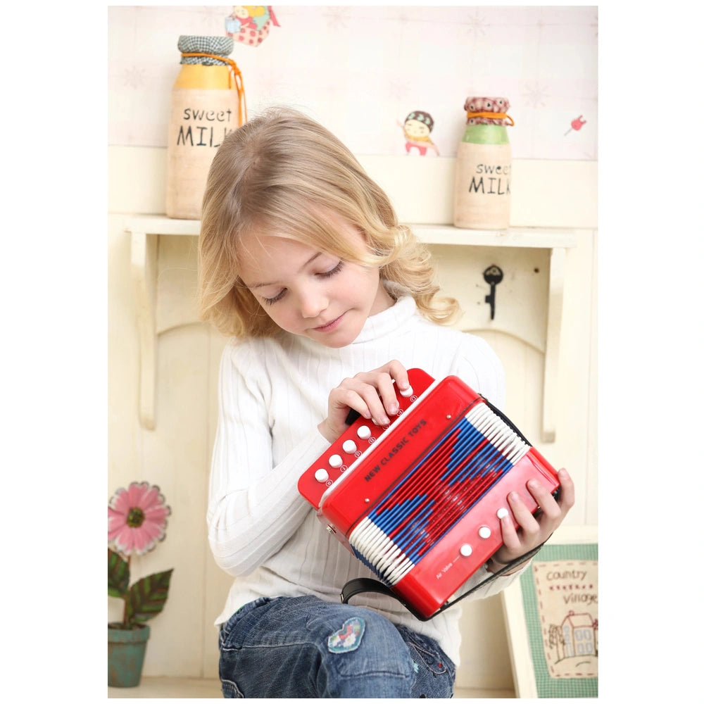 Blue accordion held by a girl in a white sweater and blue jeans, seated on a wooden bench with a floral wall behind.