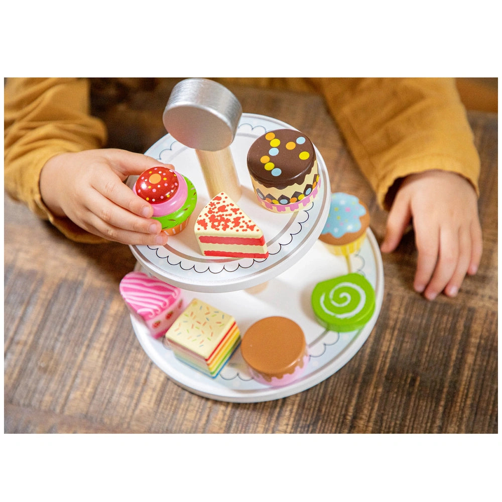 New Classic Toys cake stand set with colorful toy cakes, a child's hand, and a metallic hammer on a wooden table.