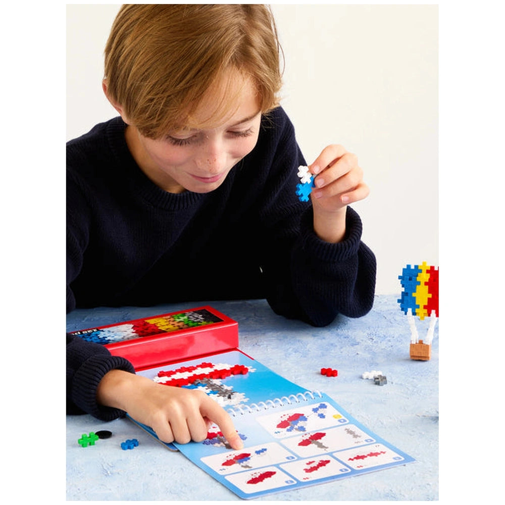 Plus Plus transportation activity pad with a child’s hand holding a glossy blue and red toy on a blue matte table.