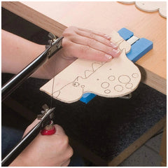 Hands using a fretsaw to cut a dinosaur shape from wood, secured with a blue clamp on a workbench.