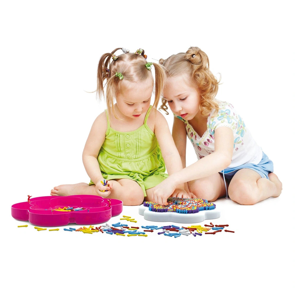 Two young girls in green tops create art with Quercetti FantaColor Mandala on a white surface surrounded by colorful beads.