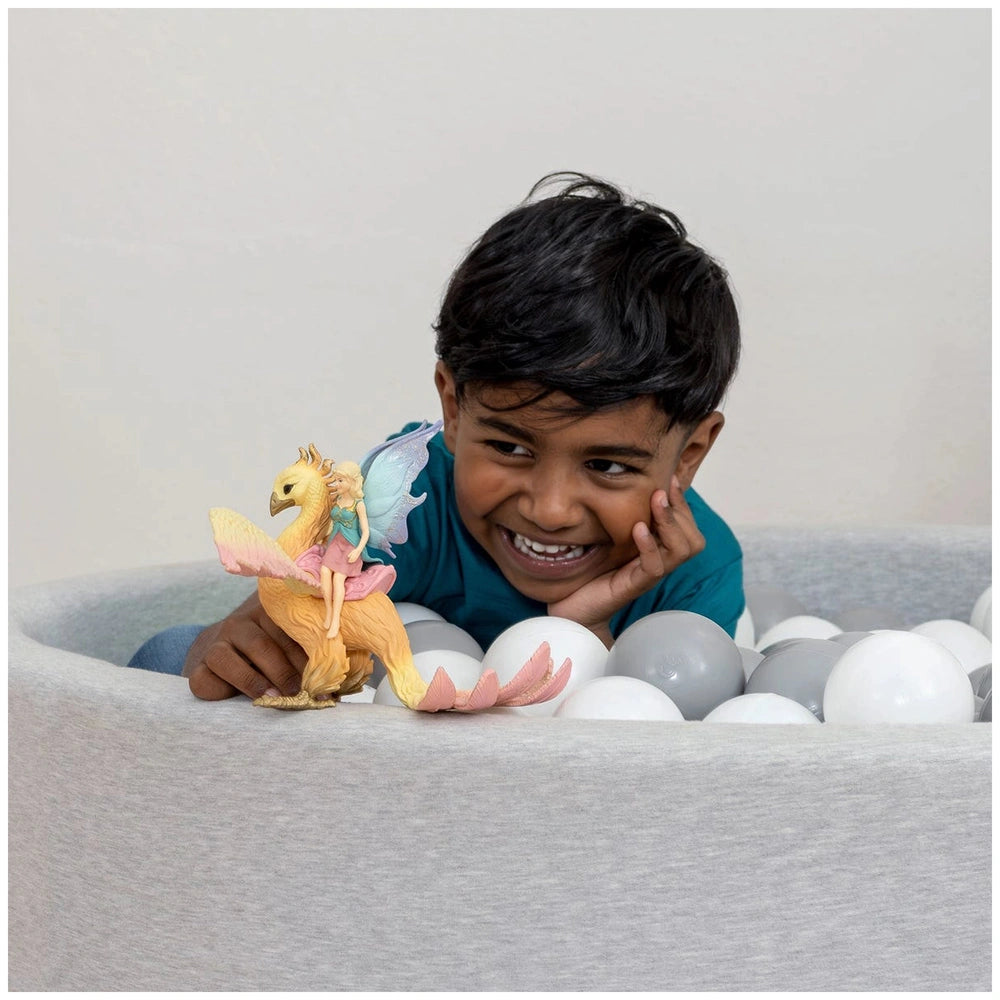 Schleich Phoenix toy held by a smiling child in a white ball pit filled with white balls, wearing a blue top.