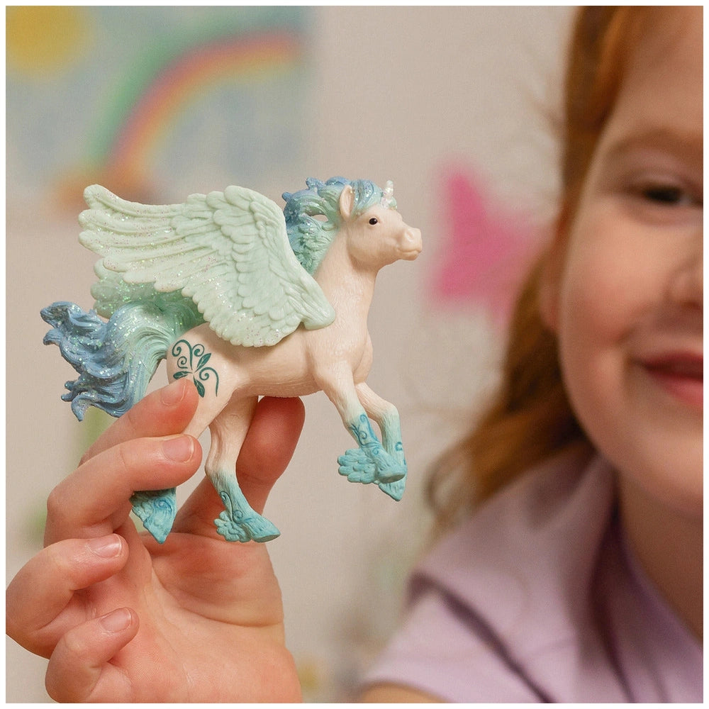 A hand holds a glossy white unicorn foal figurine with blue accents, set against a colorful, blurred indoor background.