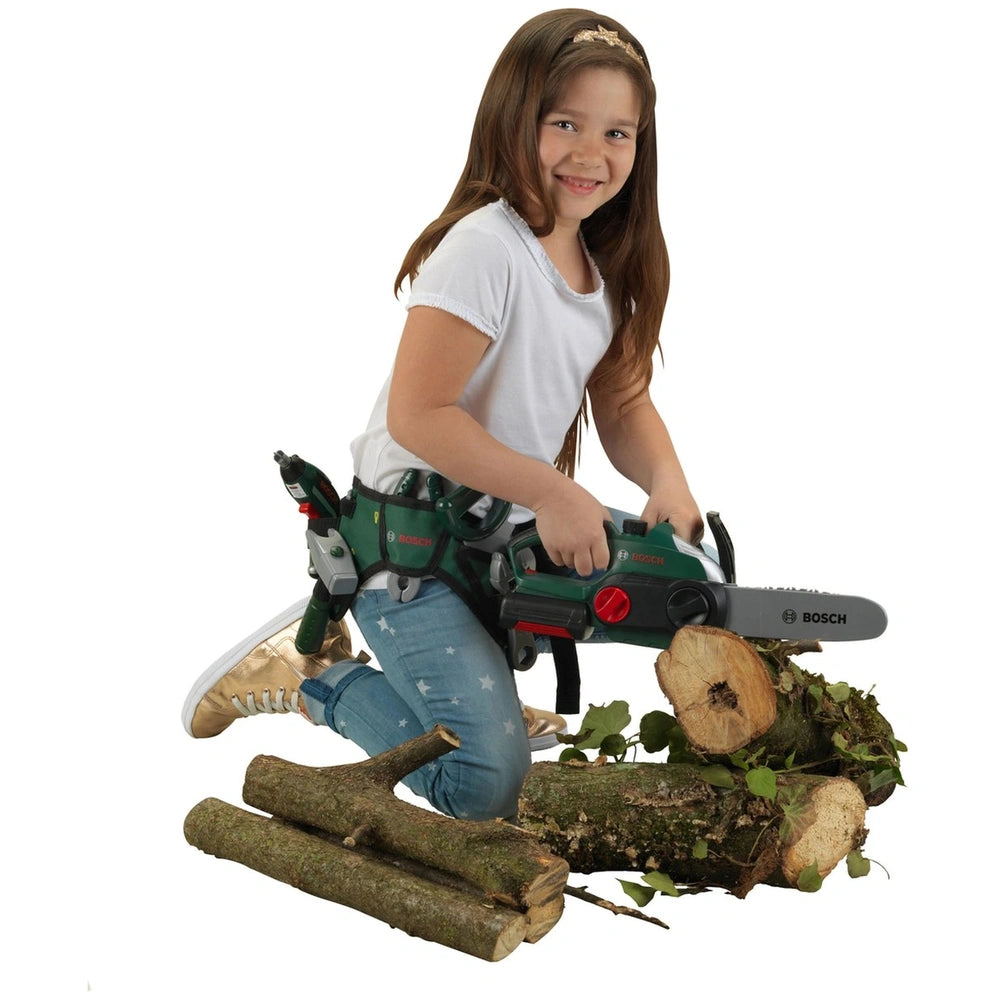 Young girl in a green safety harness holds a green chainsaw, standing on a moss-covered log in blue jeans and gold sneakers.