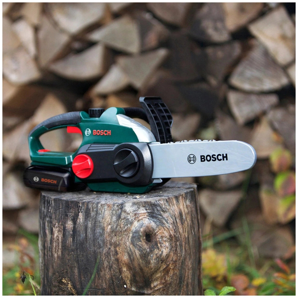 Theo Klein Bosch chain saw with green and black body, metallic blade, and black plastic handle, placed on a rough wooden stum