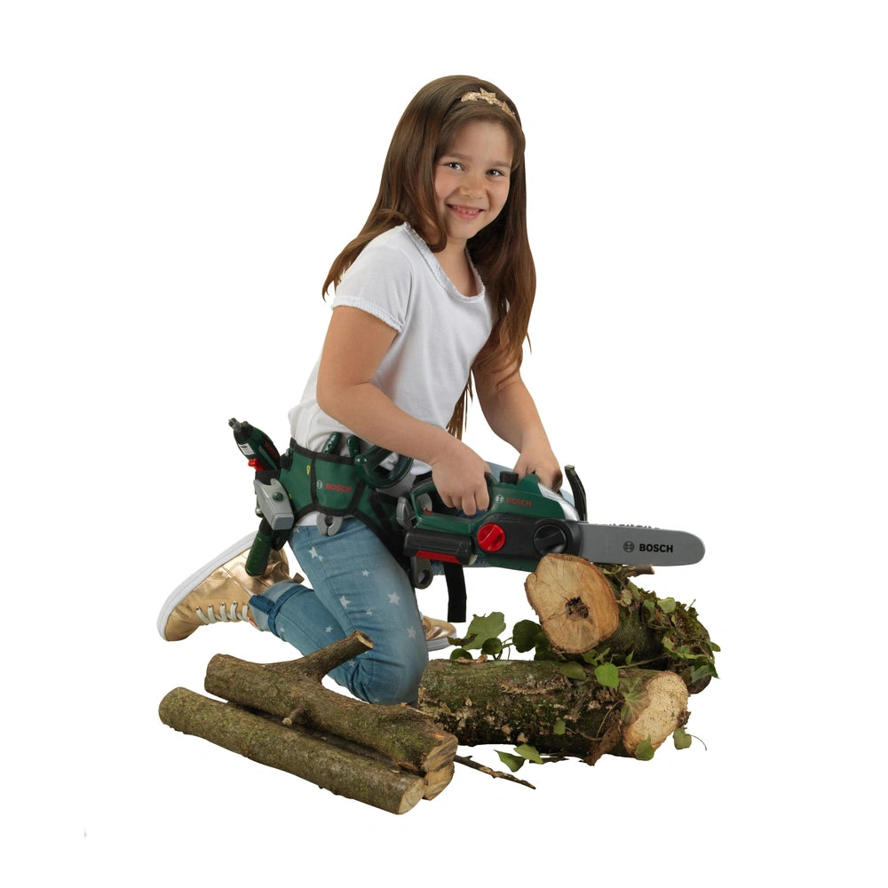 Theo Klein Bosch chainsaw toy held by a girl in a white t-shirt and blue jeans, standing on mossy logs outdoors.