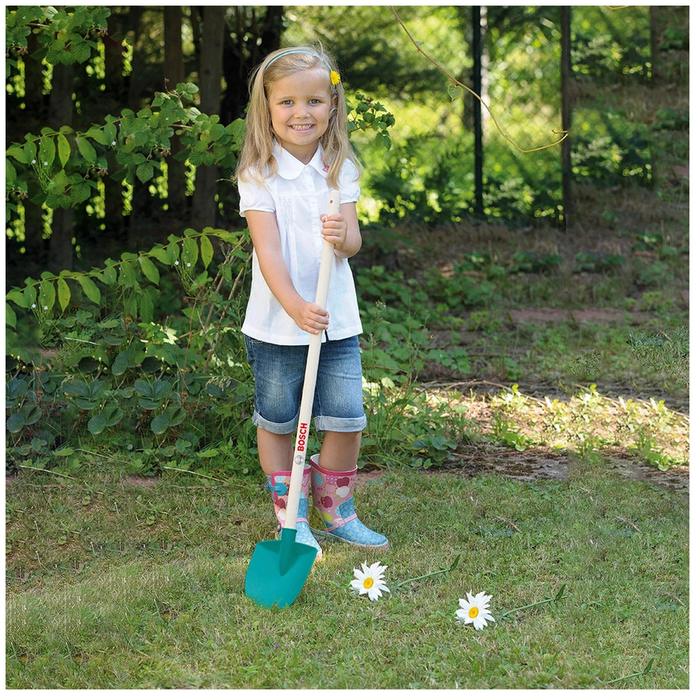 Young girl in a white shirt and blue jeans holds a green and white shovel, standing on grass with white flowers and green fol