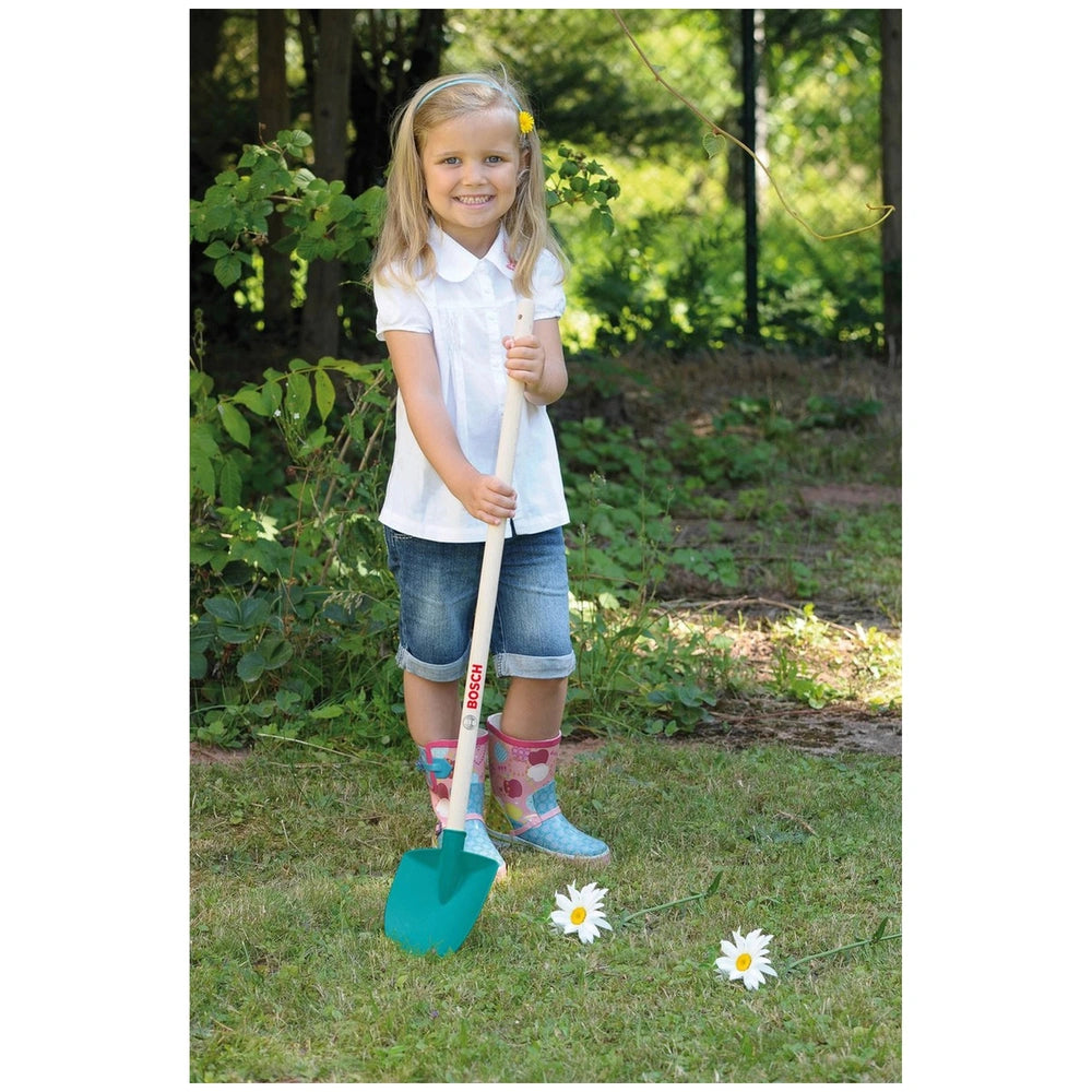 Young girl in a white shirt and blue rain boots holds a green and white shovel in a grassy area with flowers and a wooden fen