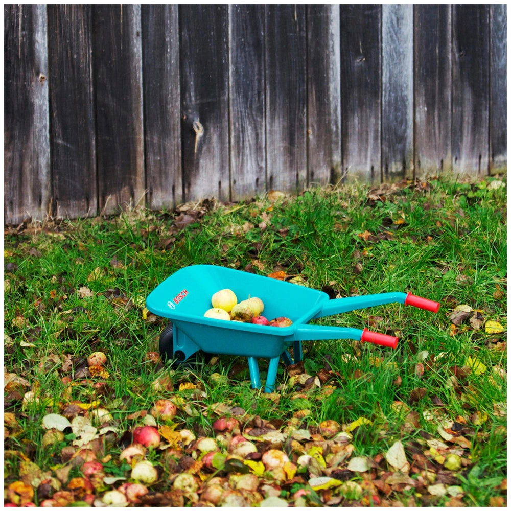 Theo Klein Bosch garden wheelbarrow in blue surrounded by green grass and red-yellow apples near a brown wooden fence.